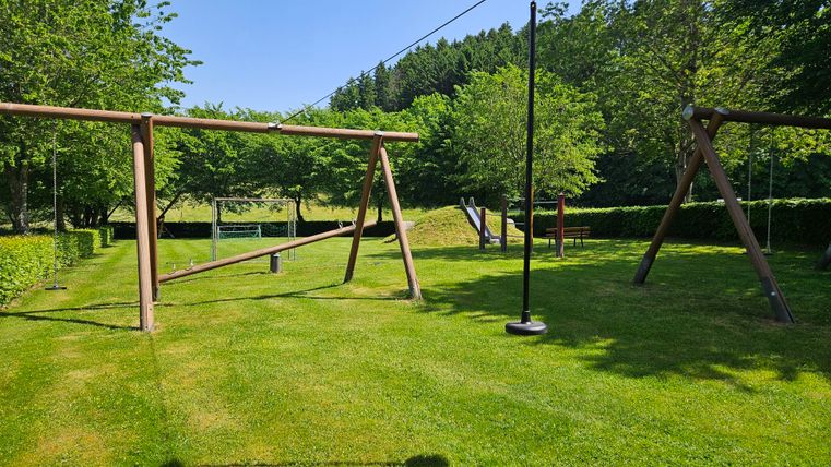 Ein Spielplatz mit Schaukeln steht auf einem grünen Rasen. Bäume umgeben den Bereich und der Himmel ist klar blau.