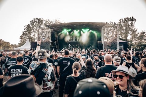 A lively concert event with a large crowd. On stage, musicians can be seen, surrounded by green lights.