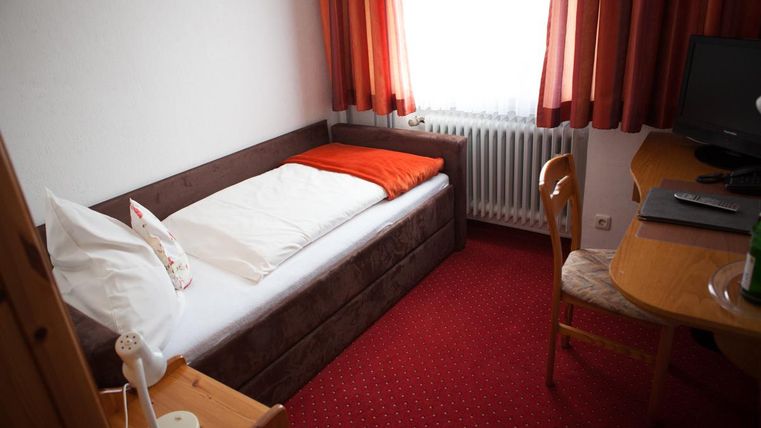 Ein einfaches Zimmer mit einem Einzelbett, einem Tisch und einem Stuhl. Rote Vorhänge lassen Tageslicht herein und schaffen eine gemütliche Atmosphäre.