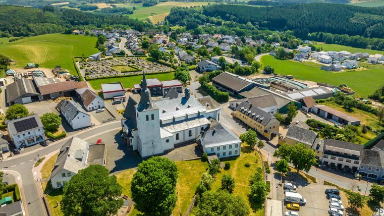 Eine idyllische Landschaft mit einer kleinen Kirche im Mittelpunkt. Im Hintergrund sind grüne Felder und Wohngebäude zu sehen.