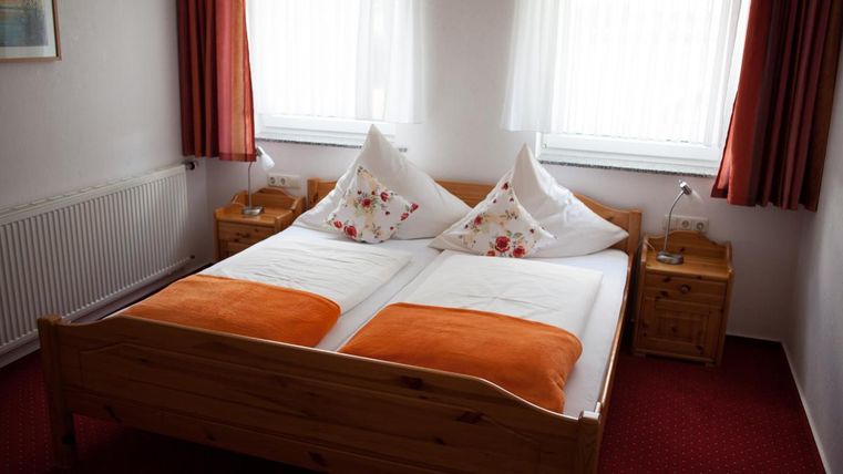 Ein gemütliches Schlafzimmer mit einem Doppelbett und orangefarbenen Decken. Die Räume sind hell und freundlich, mit Fenstern, die viel Tageslicht hereinlassen.