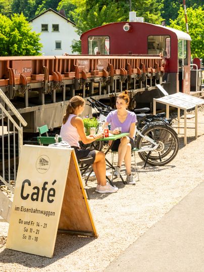 Een café in een treinstel met twee vrouwen die aan een tafel zitten. Op de achtergrond zijn fietsen en een zonnig landschap te zien.