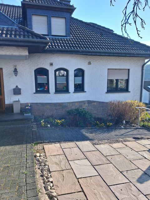 Ein modernes Wohnhaus mit einem gepflasterten Weg und einem kleinen Garten. Die Fenster sind mit Jalousien versehen und die Fassade ist hell gestrichen.