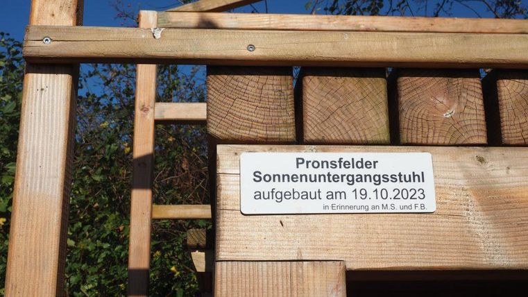 Ein Holzstuhl mit dem Aufschrift "Pronsfelder Sonnenuntergangsstuhl", der am 19.10.2023 aufgebaut wurde. Im Hintergrund sind Bäume und ein blauer Himmel zu sehen.