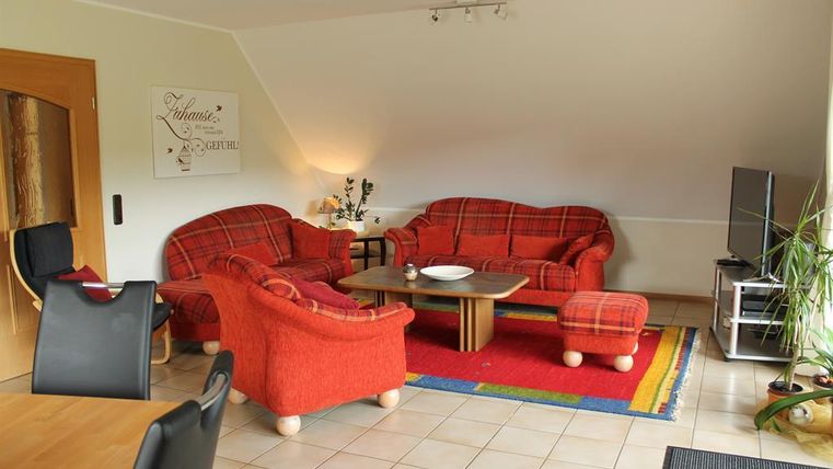 Ein gemütliches Wohnzimmer mit roten Stoffsofas und einem Holztisch. Im Hintergrund steht ein Fernseher und eine Pflanze sorgt für eine frische Atmosphäre.