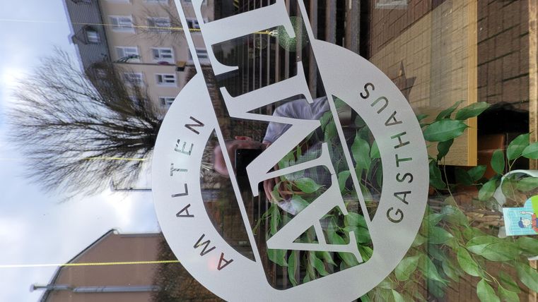 Een venster met het logo van het gasthuis "Am Alten". Op de achtergrond zijn planten en een straat zichtbaar.