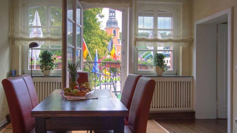 Ein gemütlicher Essbereich mit einem Holztisch und roten Stühlen. Blick aus dem Fenster auf eine Straße mit Flaggen und einem Kirchturm im Hintergrund.