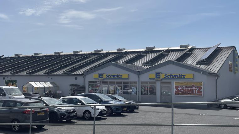 Ein Geschäftshaus mit dem Namen "Schmitz" und einem modernen Dach. Vor dem Gebäude parken mehrere Autos auf einem großen Parkplatz.