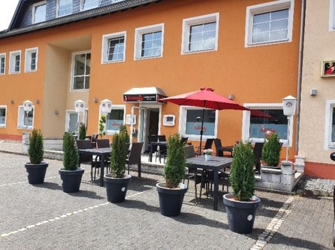 Ein modernes Gebäude mit orangefarbener Fassade und großen Fenstern. Draußen stehen Tische mit Stühlen und ein roter Sonnenschirm, umgeben von Pflanzen in schwarz-weißen Töpfen.
