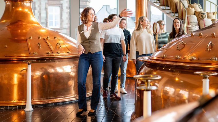 Een groep mensen bezoekt een brouwerij. Een vrouw legt iets uit bij grote koperen ketels.