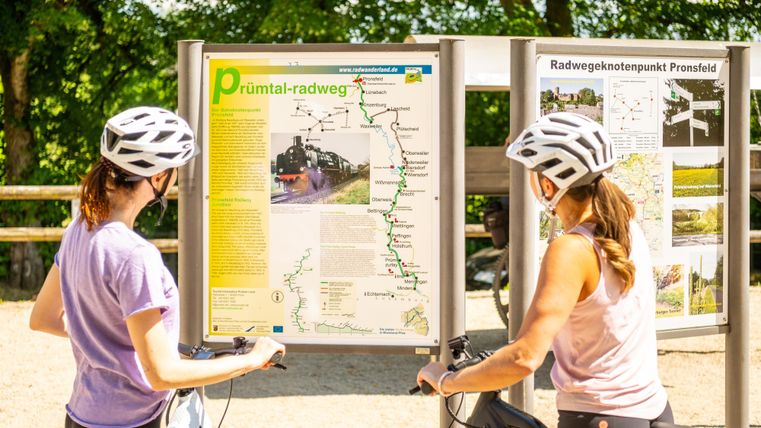 Zwei Radfahrerinnen stehen vor einem Informationsschild über den Prüm-Tal-Radweg. Sie tragen Helme und schauen sich die Karte und Informationen an.