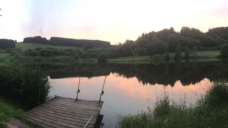 Ein ruhiger Teich mit einem Holzsteg und üppigem Grün. Der Himmel ist in sanften Farben bei Sonnenuntergang gefärbt.