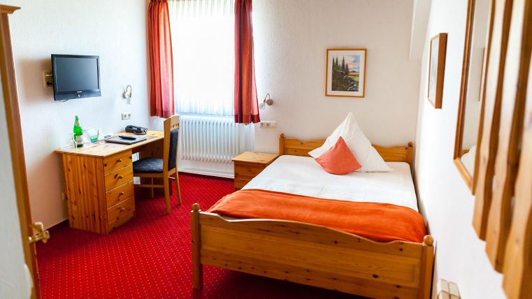 Ein gemütliches Hotelzimmer mit einem Einzelbett und einem orangefarbenen Bettüberwurf. Am Tisch stehen ein Stuhl und eine Flasche Wasser, und ein Fernseher hängt an der Wand.