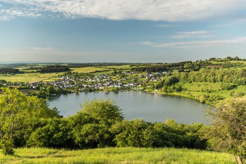 Eine malerische Landschaft mit einem ruhigen See und einem kleinen Dorf im Hintergrund. Die Umgebung ist grün und von sanften Hügeln umgeben.